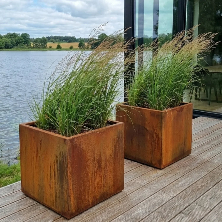 Steelart CUBE kubo formos metaliniai lauko vazonai natūralios plieno spalvos medinėje terasoje prie ežero